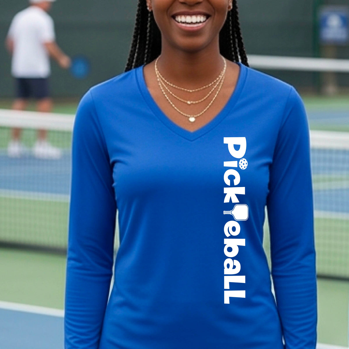 A woman wearing a royal blue long-sleeve t-shirt with a white graphic text 'PICKLEBALL' with a pickleball paddle replacing the L, on the front, standing on a pickleball court.