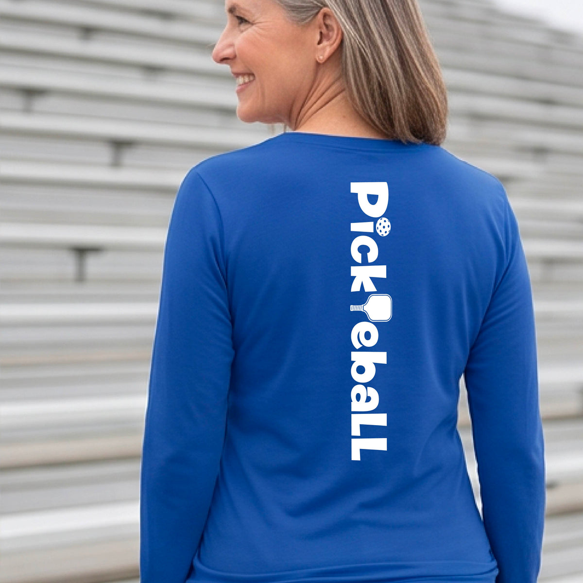 A woman wearing a royal blue long-sleeve t-shirt with a white graphic text 'PICKLEBALL' with a pickleball paddle replacing the L, on the back, standing on a pickleball court.