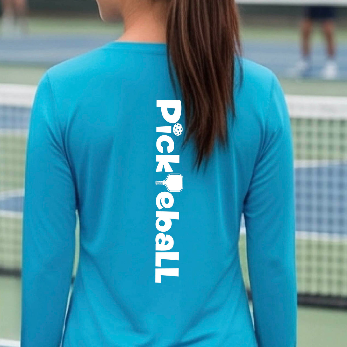 A woman wearing a bright blue long-sleeve t-shirt with a white graphic text 'PICKLEBALL' with a pickleball paddle replacing the L, on the back, standing on a pickleball court.