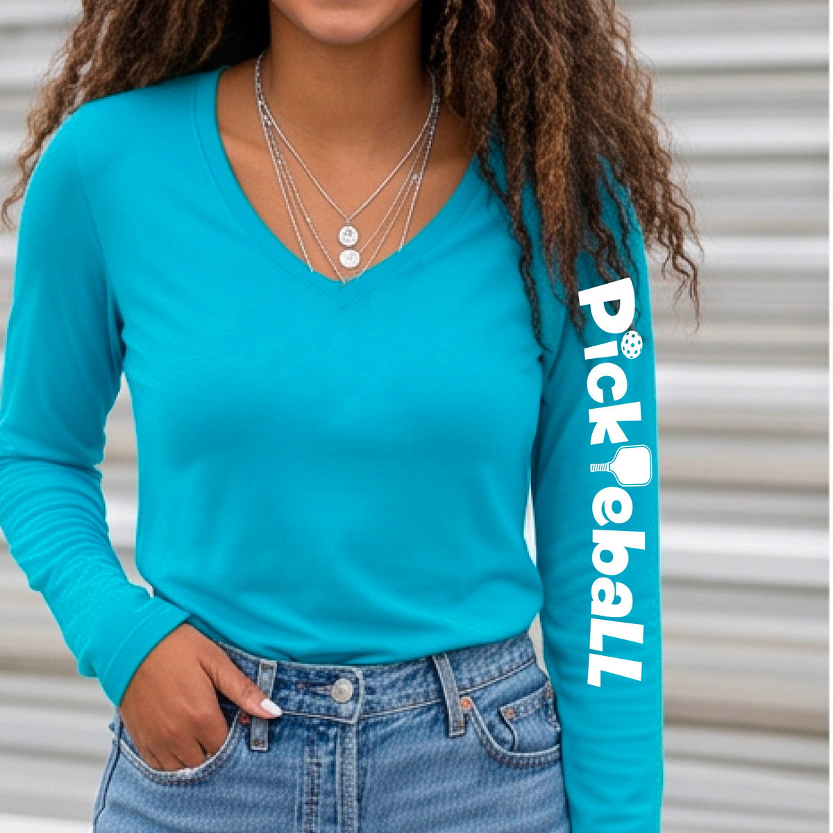 A woman wearing a bright blue long-sleeve t-shirt with a white graphic text 'PICKLEBALL' with a pickleball paddle replacing the L, on the sleeve, standing on a pickleball court.