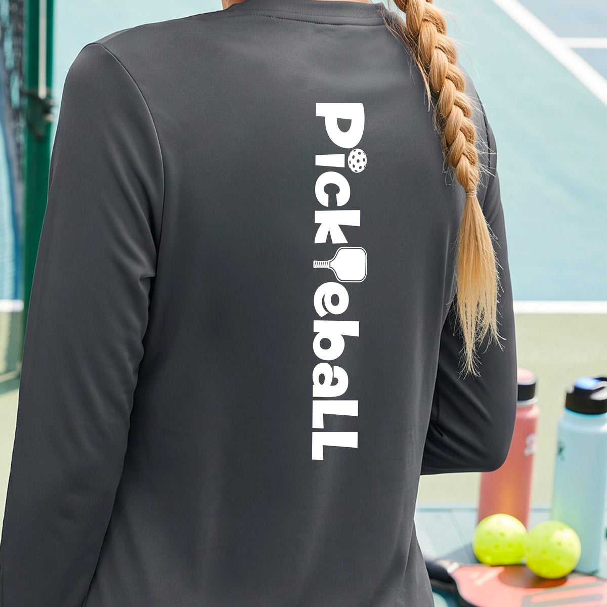 A woman wearing a gray long-sleeve t-shirt with a white graphic text 'PICKLEBALL' with a pickleball paddle replacing the L, on the back, standing on a pickleball court.