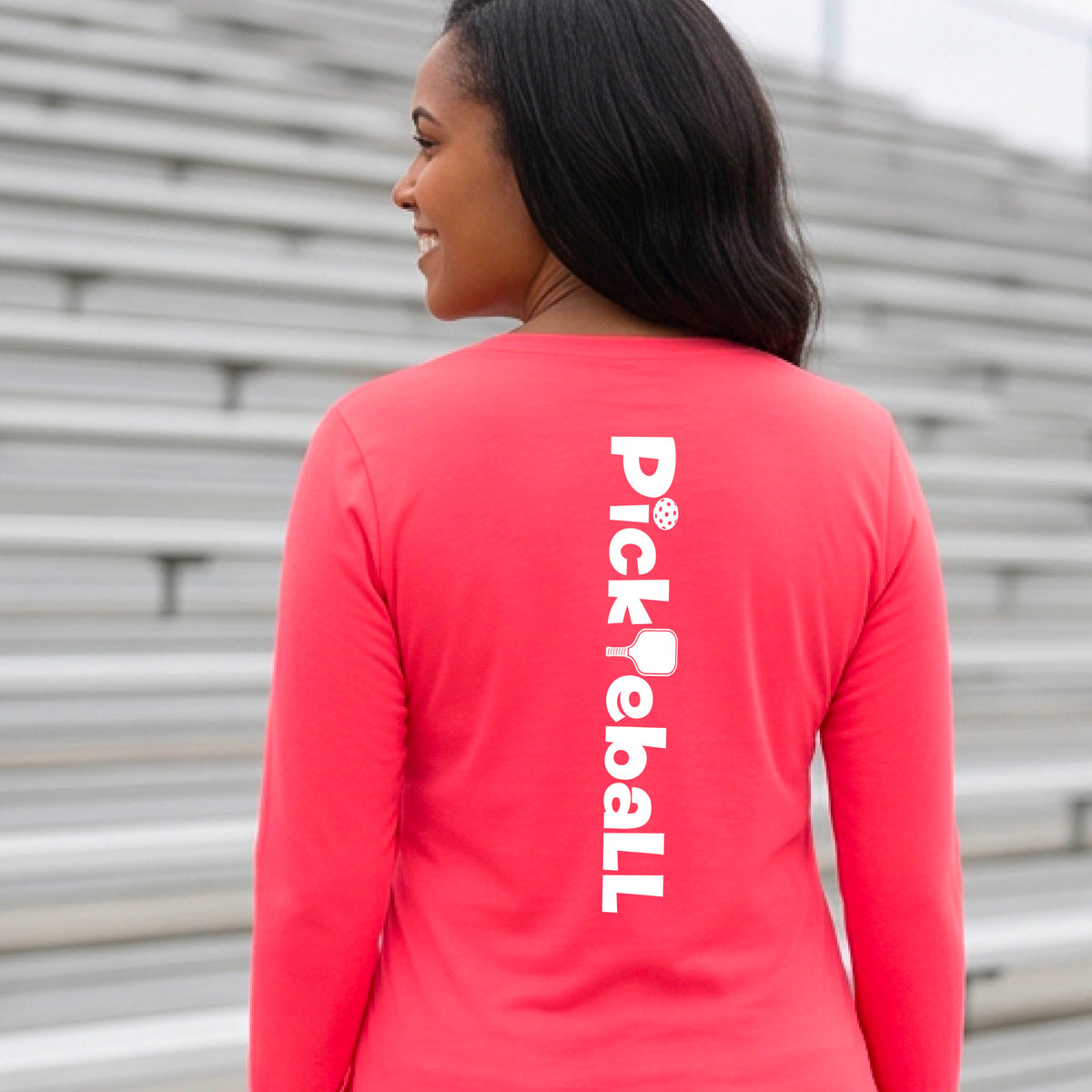 A woman wearing a coral pink long-sleeve t-shirt with a white graphic text 'PICKLEBALL' with a pickleball paddle replacing the L, on the back, standing on a pickleball court.