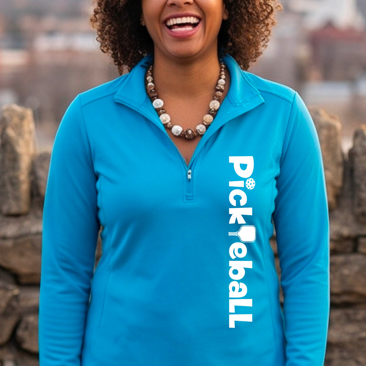 A woman wearing a bright blue quarter-zip shirt with a white graphic text 'PICKLEBALL' with a pickleball paddle replacing the L, on the front, standing on a pickleball court.