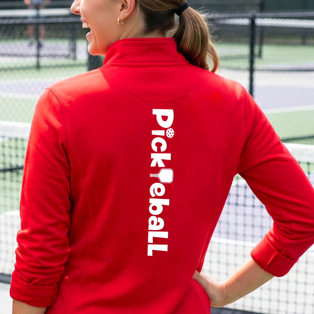A woman wearing a red quarter-zip shirt with a white graphic text 'PICKLEBALL' with a pickleball paddle replacing the L, on the back, standing on a pickleball court.