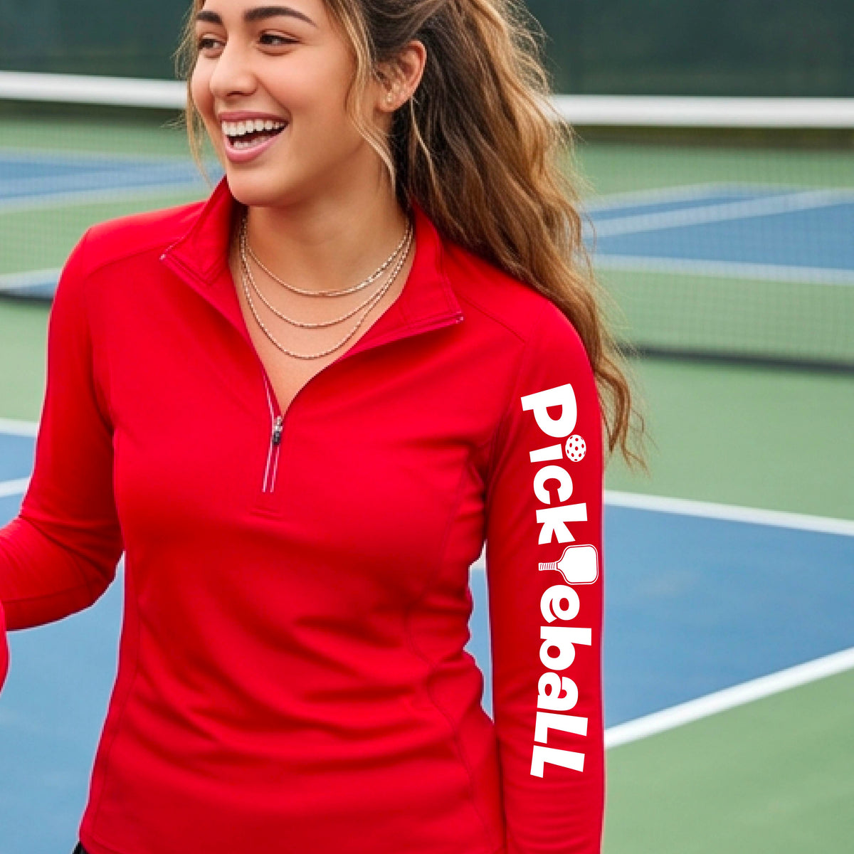 A woman wearing a red quarter-zip shirt with a white graphic text 'PICKLEBALL' with a pickleball paddle replacing the L, on the sleeve, standing on a pickleball court.