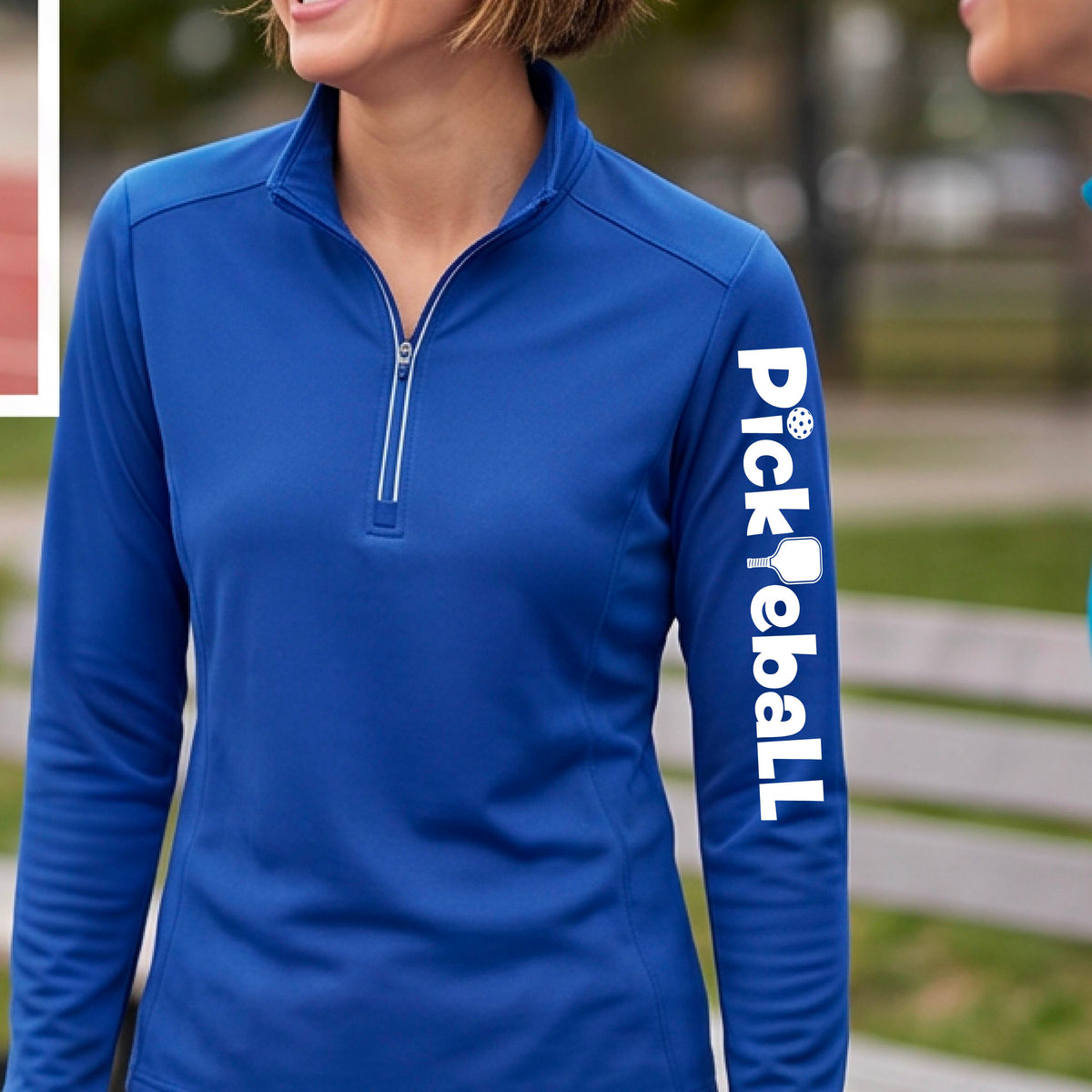 A woman wearing a royal blue quarter-zip shirt with a white graphic text 'PICKLEBALL' with a pickleball paddle replacing the L, on the sleeve, standing on a pickleball court.