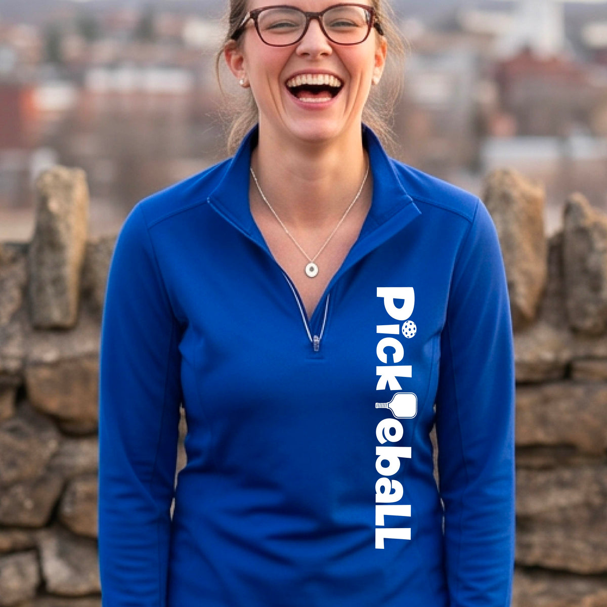 A woman wearing a royal blue quarter-zip shirt with a white graphic text 'PICKLEBALL' with a pickleball paddle replacing the L, on the front, standing on a pickleball court.