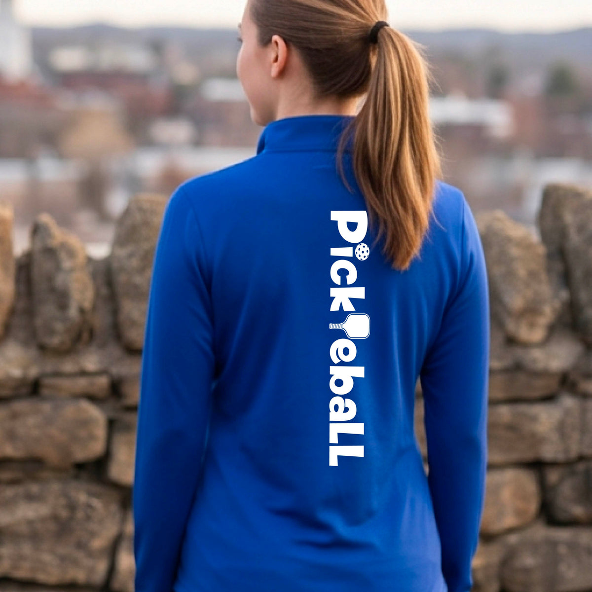 A woman wearing a royal blue quarter-zip shirt with a white graphic text 'PICKLEBALL' with a pickleball paddle replacing the L, on the back, standing on a pickleball court.