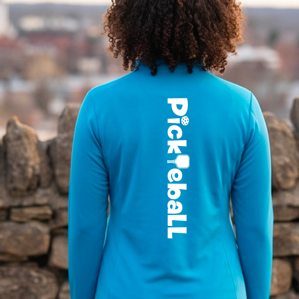 A woman wearing a bright blue quarter-zip shirt with a white graphic text 'PICKLEBALL' with a pickleball paddle replacing the L, on the back, standing on a pickleball court.