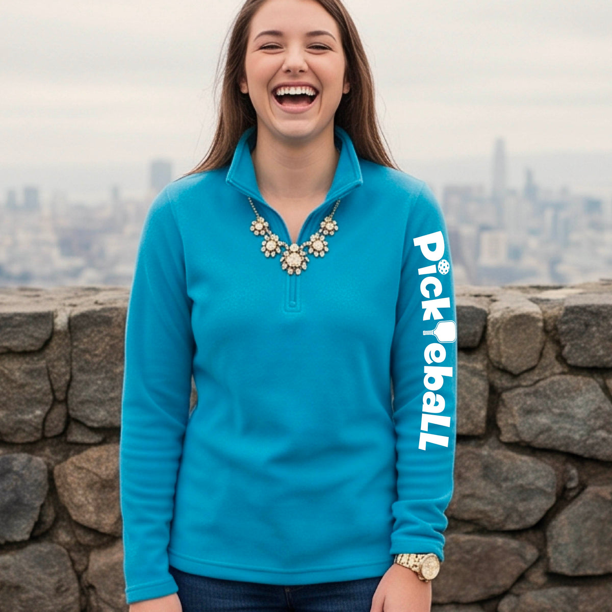 A woman wearing a bright blue quarter-zip shirt with a white graphic text 'PICKLEBALL' with a pickleball paddle replacing the L, on the sleeve, standing on a pickleball court.