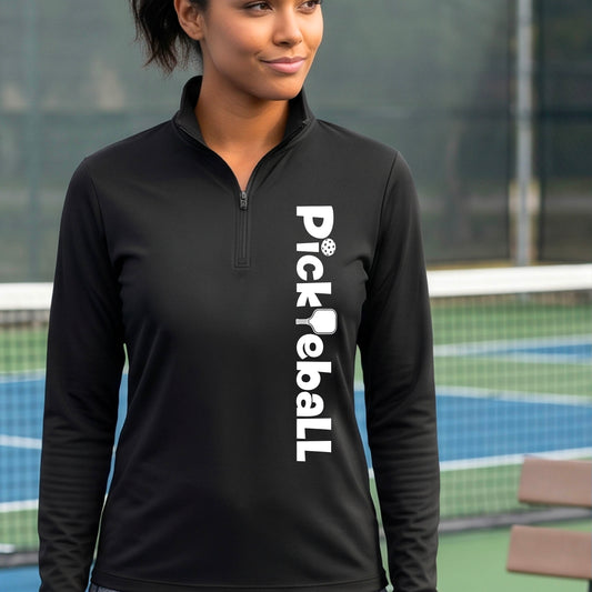 A woman wearing a black quarter-zip shirt with a white graphic text 'PICKLEBALL' with a pickleball paddle replacing the L, on the front, standing on a pickleball court.
