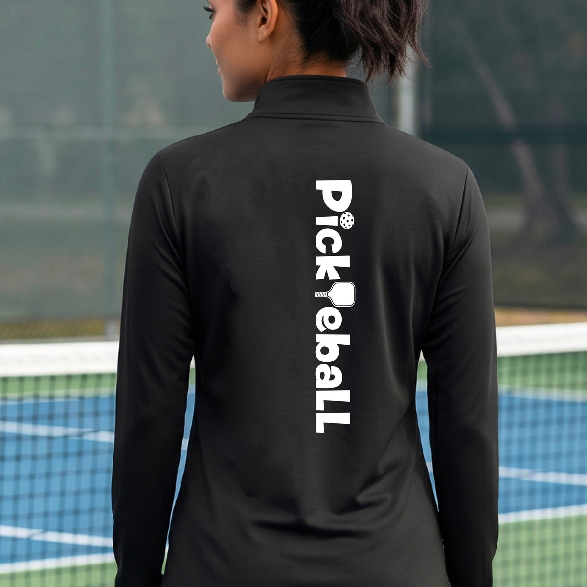 A woman wearing a black quarter-zip shirt with a white graphic text 'PICKLEBALL' with a pickleball paddle replacing the L, on the black, standing on a pickleball court.