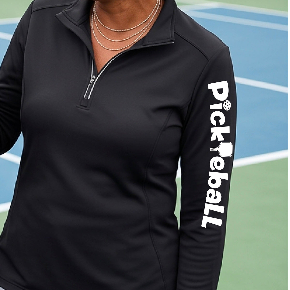 A woman wearing a quarter-zip shirt with a white graphic text 'PICKLEBALL' with a pickleball paddle replacing the L, on the sleeve, standing on a pickleball court.