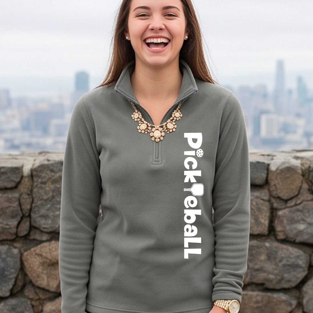A woman wearing a gray quarter-zip shirt with a white graphic text 'PICKLEBALL' with a pickleball paddle replacing the L, on the front, standing on a pickleball court.