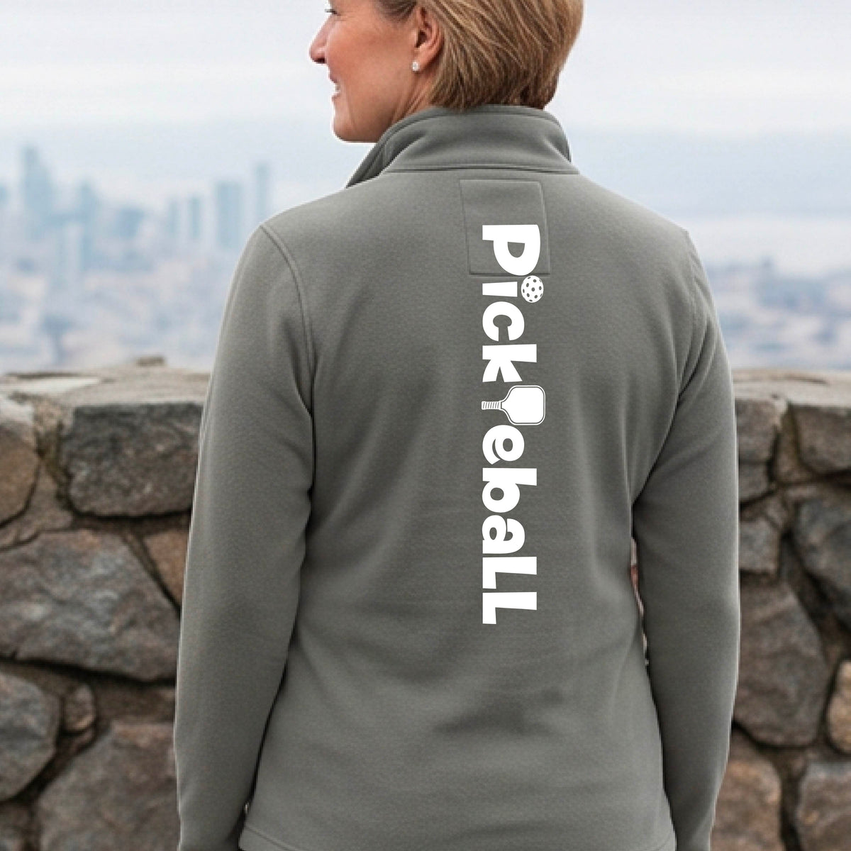 A woman wearing a gray quarter-zip shirt with a white graphic text 'PICKLEBALL' with a pickleball paddle replacing the L, on the back, standing on a pickleball court.
