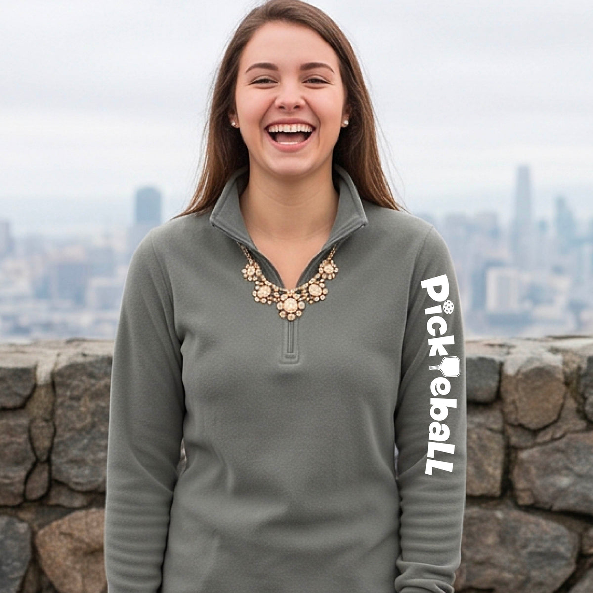 A woman wearing a gray quarter-zip shirt with a white graphic text 'PICKLEBALL' with a pickleball paddle replacing the L, on the sleeve standing on a pickleball court.