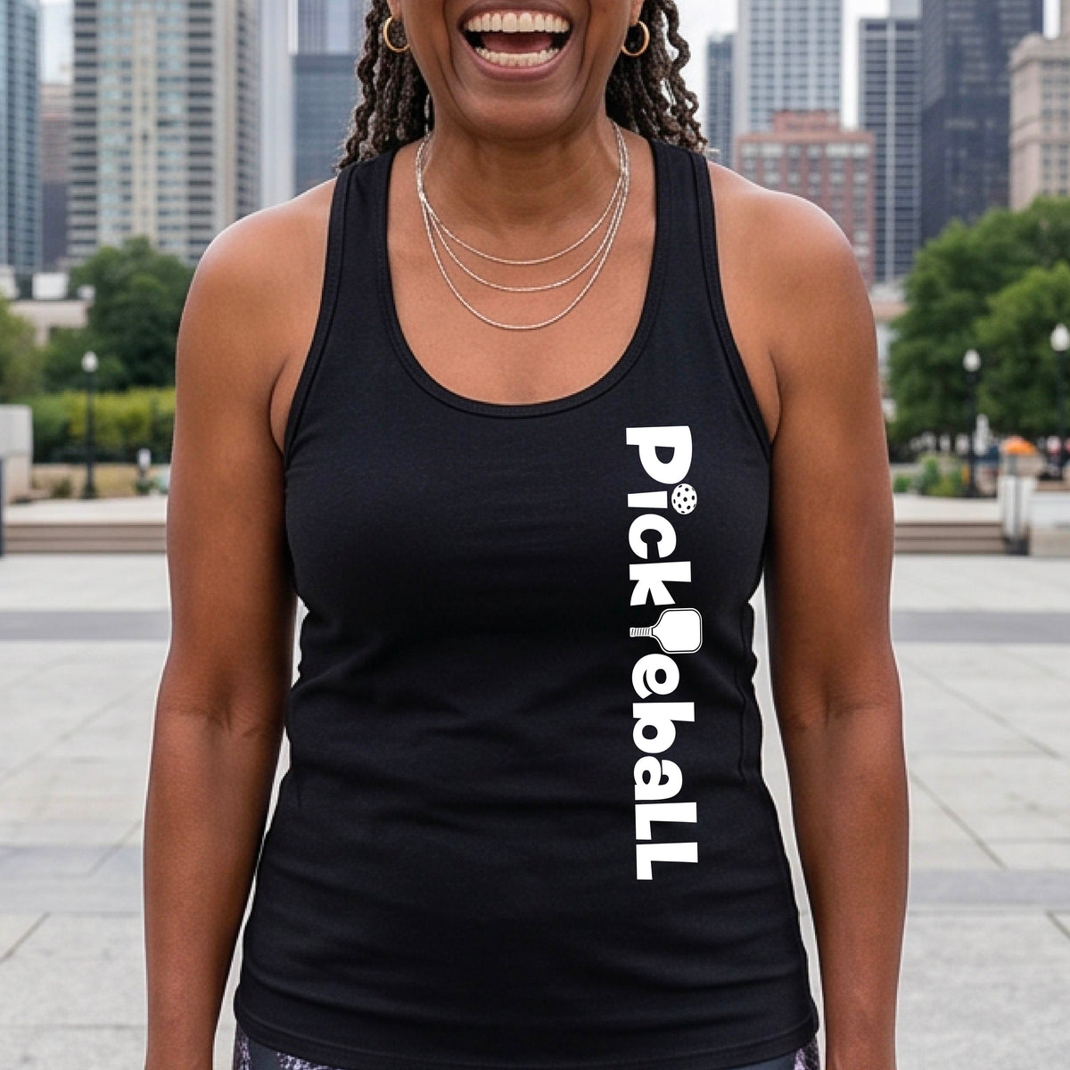 A woman wearing a black racerback tank top with a white graphic text 'PICKLEBALL' with a pickleball paddle replacing the L, on the front, standing on a pickleball court.