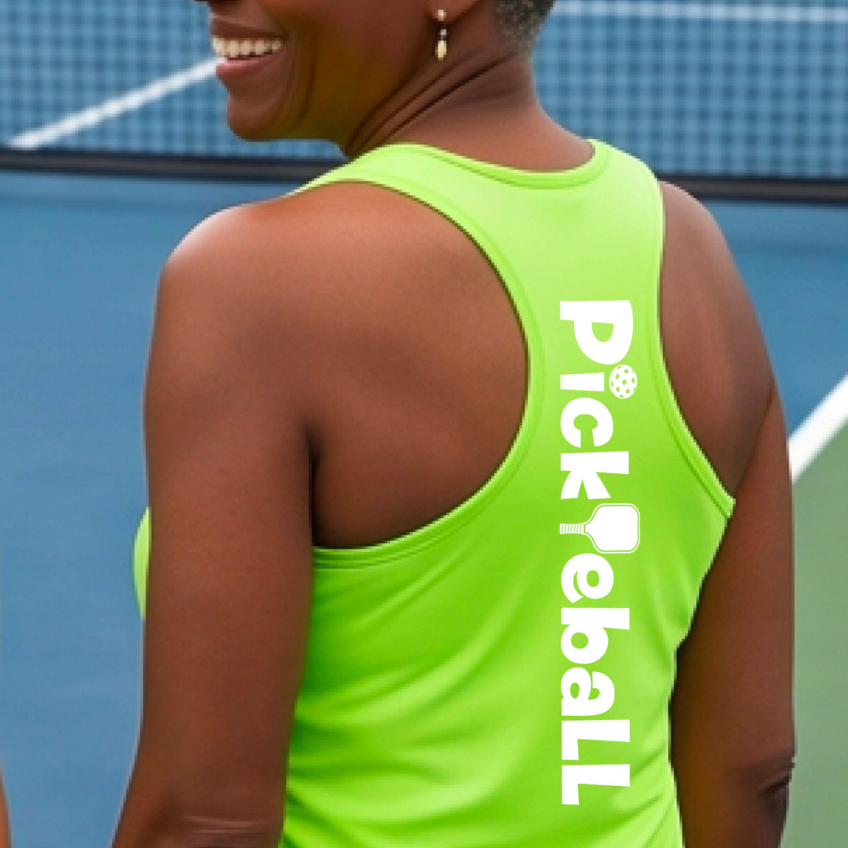 A woman wearing a lime green racerback tank top with a white graphic text 'PICKLEBALL' with a pickleball paddle replacing the L, on the back, standing on a pickleball court.