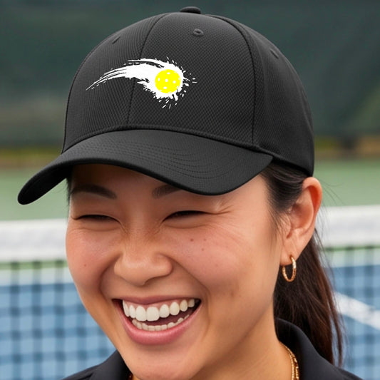 A close-up of a black pickleball hat with a white pickleball graphic of a yellow pickleball with a motion trail effect, displayed in front of a pickleball court. 