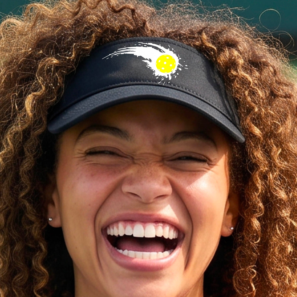 A close-up of a black visor with a white pickleball graphic of a yellow pickleball with a motion trail effect, displayed in front of a pickleball court. 