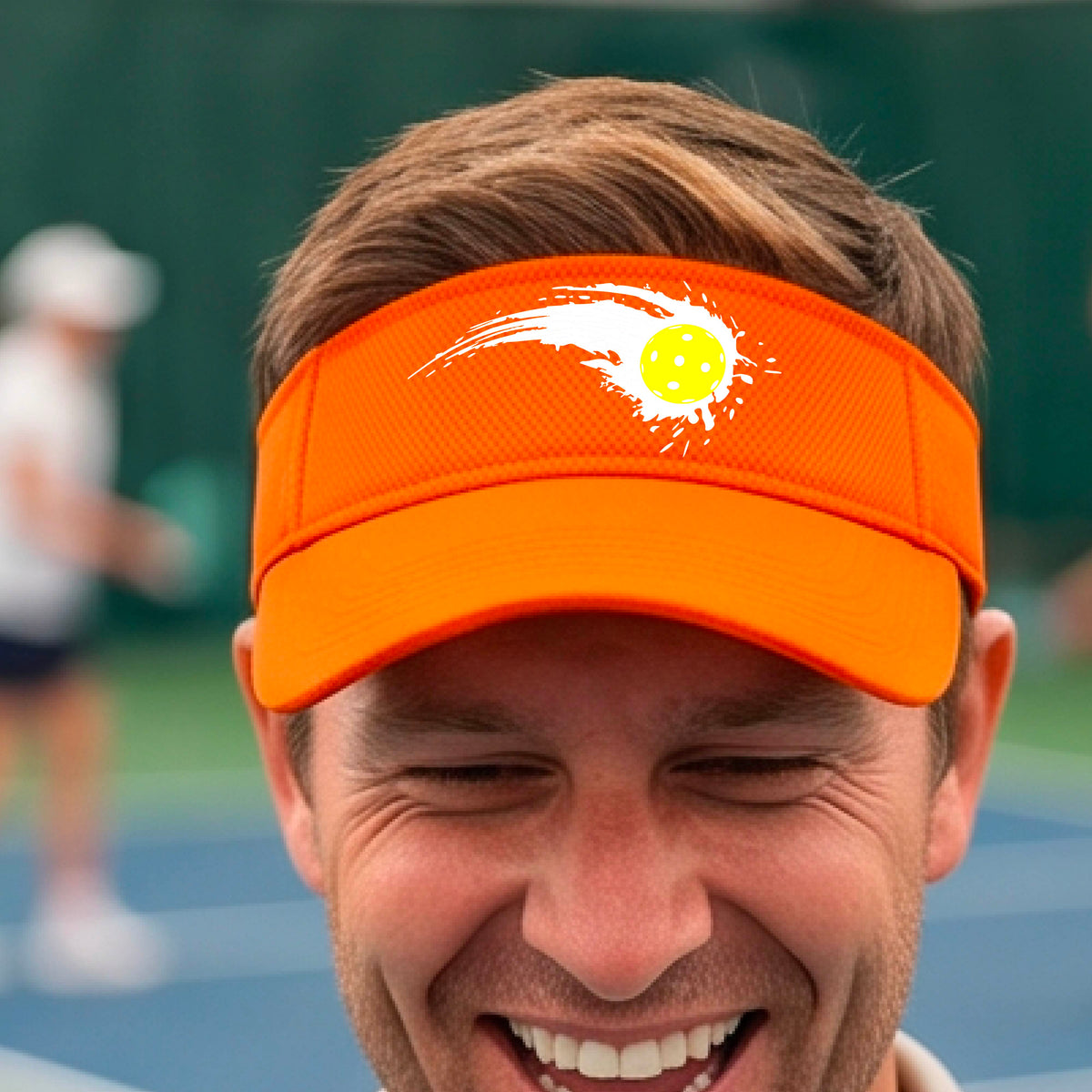 A close-up of an orange visor with a white pickleball graphic of a yellow pickleball with a motion trail effect, displayed in front of a pickleball court. 