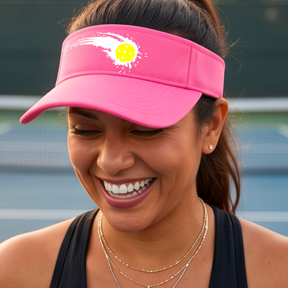 A close-up of a pink visor with a white pickleball graphic of a yellow pickleball with a motion trail effect, displayed in front of a pickleball court. 