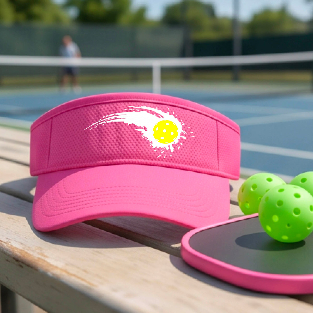 A close-up of a pink visor with a white pickleball graphic of a yellow pickleball with a motion trail effect, displayed in front of a pickleball court. 