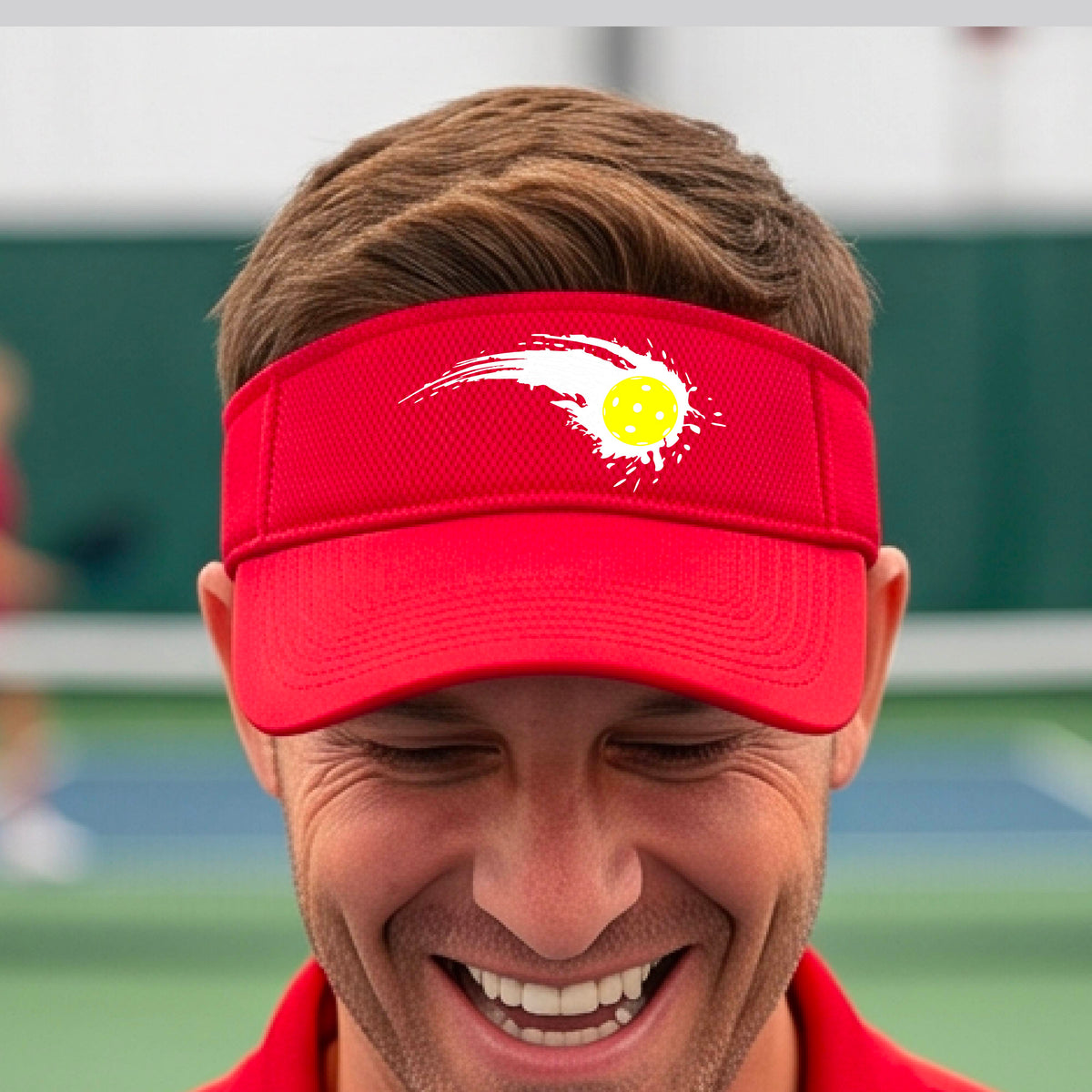A close-up of a red visor with a white pickleball graphic of a yellow pickleball with a motion trail effect, displayed in front of a pickleball court. 