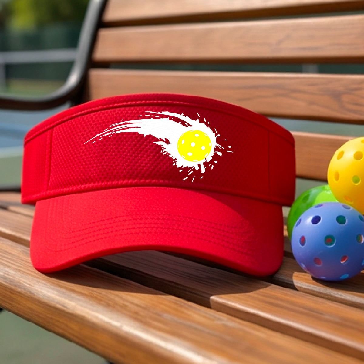 A close-up of a red visor with a white pickleball graphic of a yellow pickleball with a motion trail effect, displayed in front of a pickleball court. 