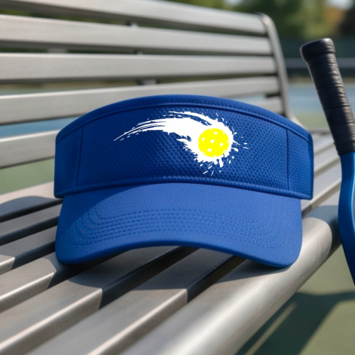 A close-up of a royal blue visor with a white pickleball graphic of a yellow pickleball with a motion trail effect, displayed in front of a pickleball court. 