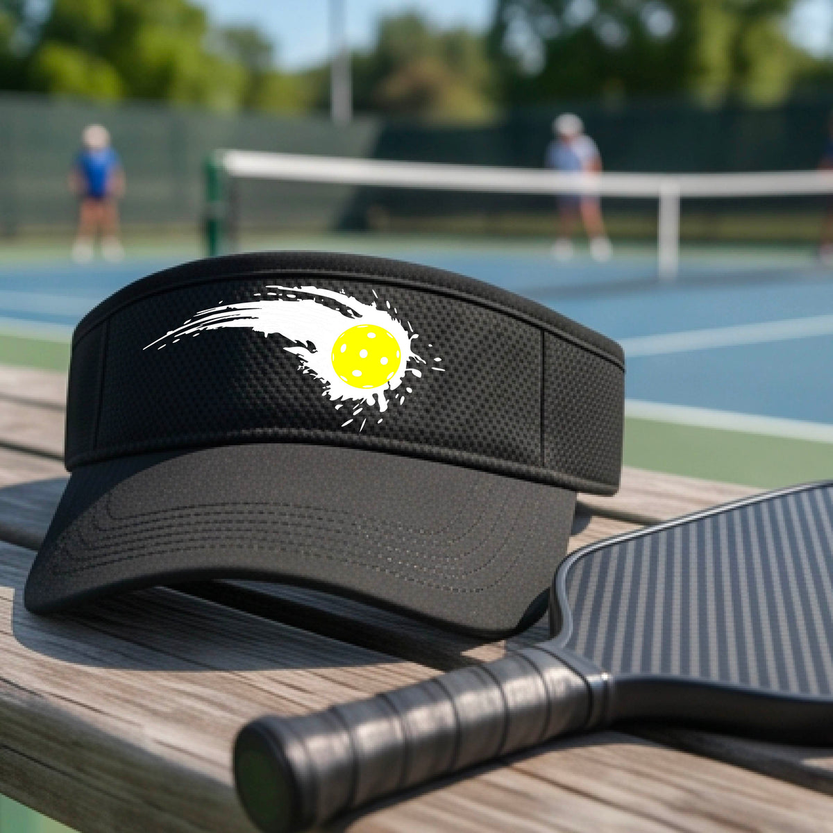 A close-up of a black visor with a white pickleball graphic of a yellow pickleball with a motion trail effect, displayed in front of a pickleball court. 