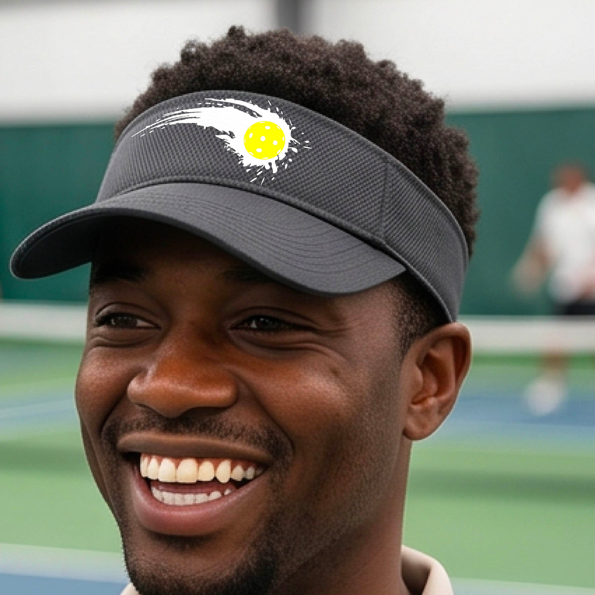 A close-up of a dark gray visor with a white pickleball graphic of a yellow pickleball with a motion trail effect, displayed in front of a pickleball court. 