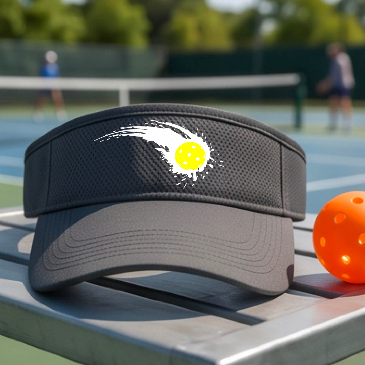 A close-up of a dark gray visor with a white pickleball graphic of a yellow pickleball with a motion trail effect, displayed in front of a pickleball court. 