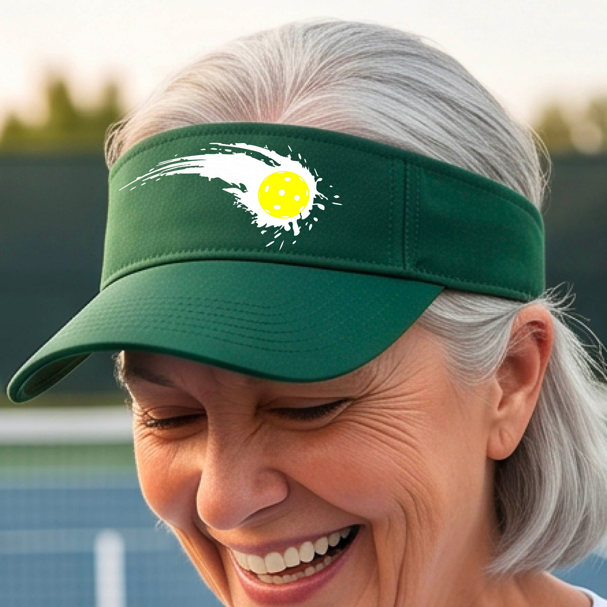 A close-up of a green visor with a white pickleball graphic of a yellow pickleball with a motion trail effect, displayed in front of a pickleball court. 
