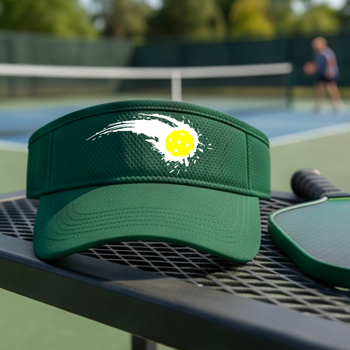 A close-up of a green visor with a white pickleball graphic of a yellow pickleball with a motion trail effect, displayed in front of a pickleball court. 