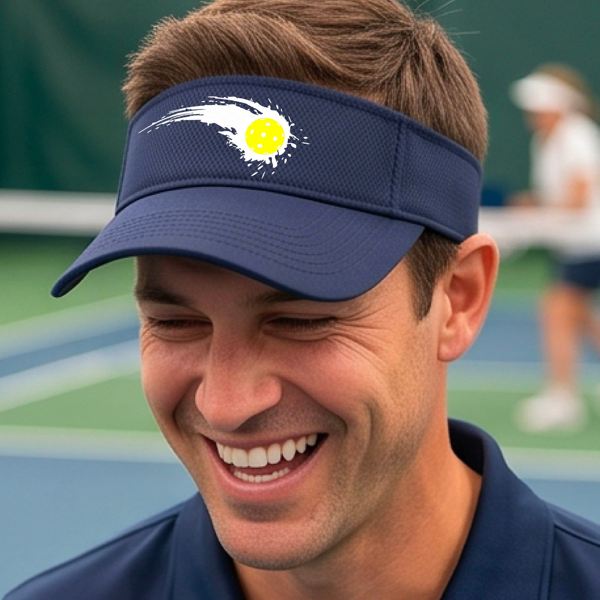 A close-up of a navy blue visor with a white pickleball graphic of a yellow pickleball with a motion trail effect, displayed in front of a pickleball court. 