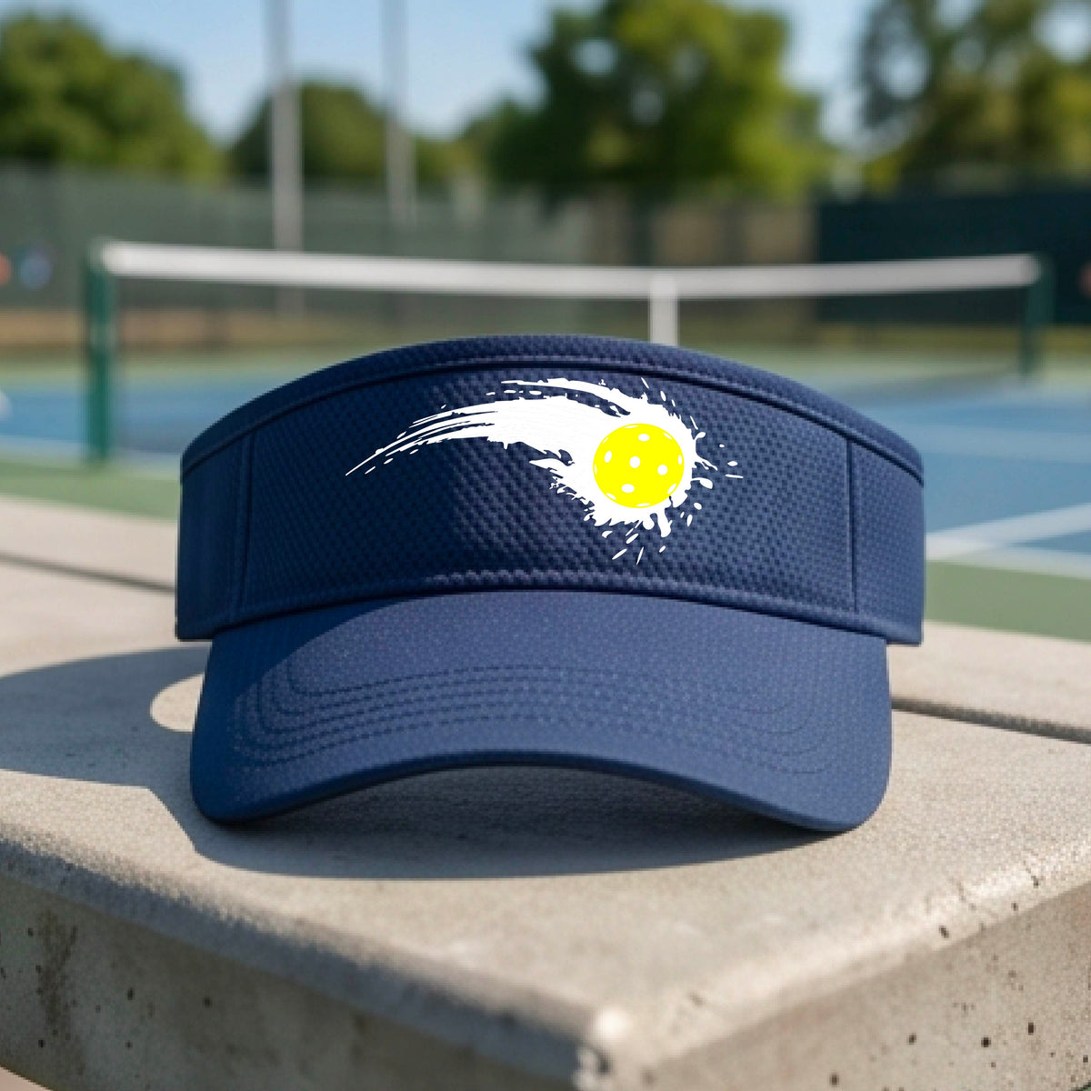 A close-up of a navy blue visor with a white pickleball graphic of a yellow pickleball with a motion trail effect, displayed in front of a pickleball court. 