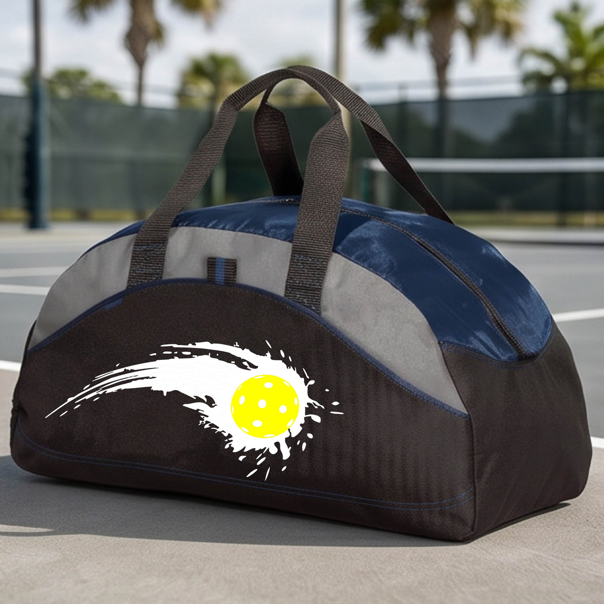 A close-up of a navy blue, black and gray medium size duffel bag with a white pickleball graphic of a yellow pickleball with a motion trail effect, displayed in front of a pickleball court. 
