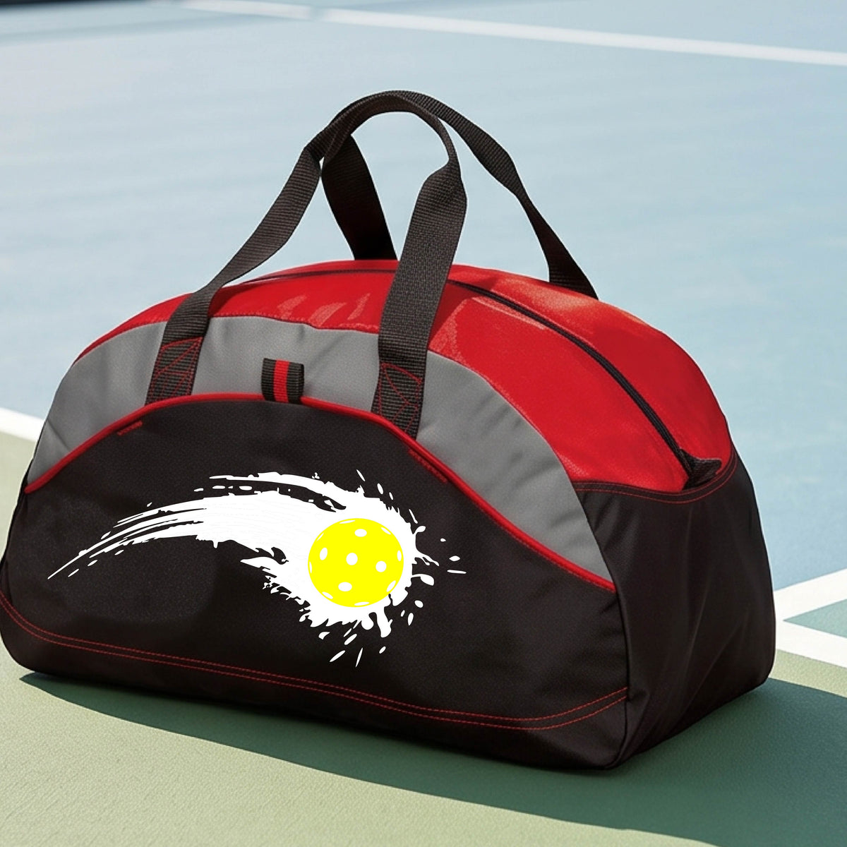 A close-up of a red, black and gray medium size duffel bag with a white pickleball graphic of a yellow pickleball with a motion trail effect, displayed in front of a pickleball court. 