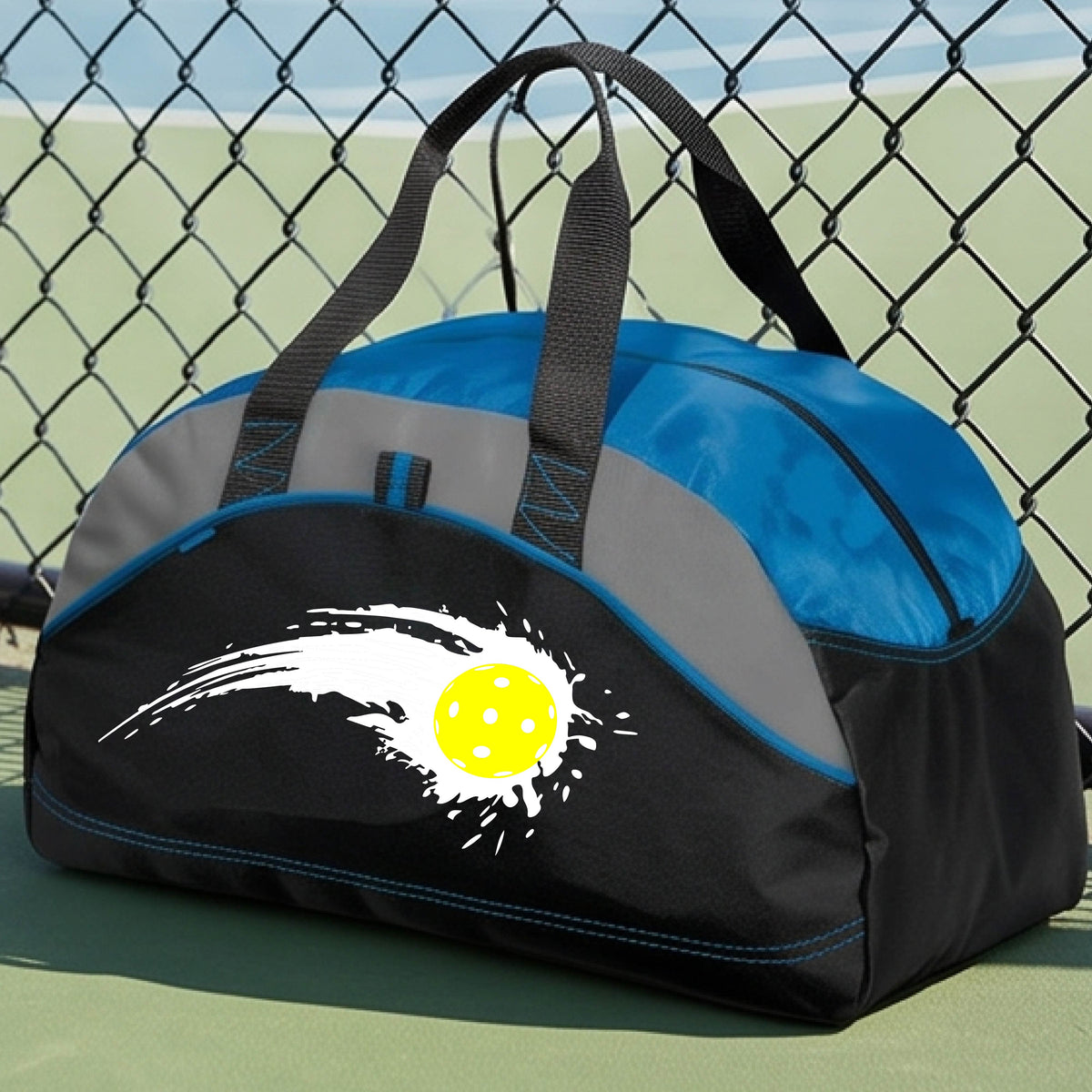 A close-up of a royal blue, black and gray medium size duffel bag with a white pickleball graphic of a yellow pickleball with a motion trail effect, displayed in front of a pickleball court. 