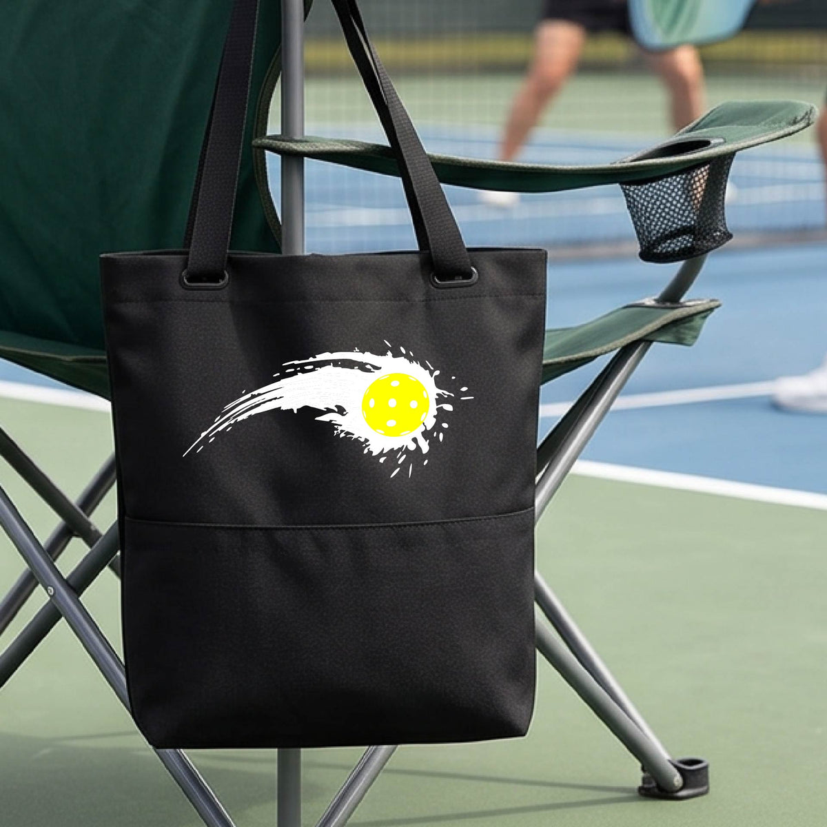 A close-up of a black tote bag with a white pickleball graphic of a yellow pickleball with a motion trail effect, displayed in front of a pickleball court. 
