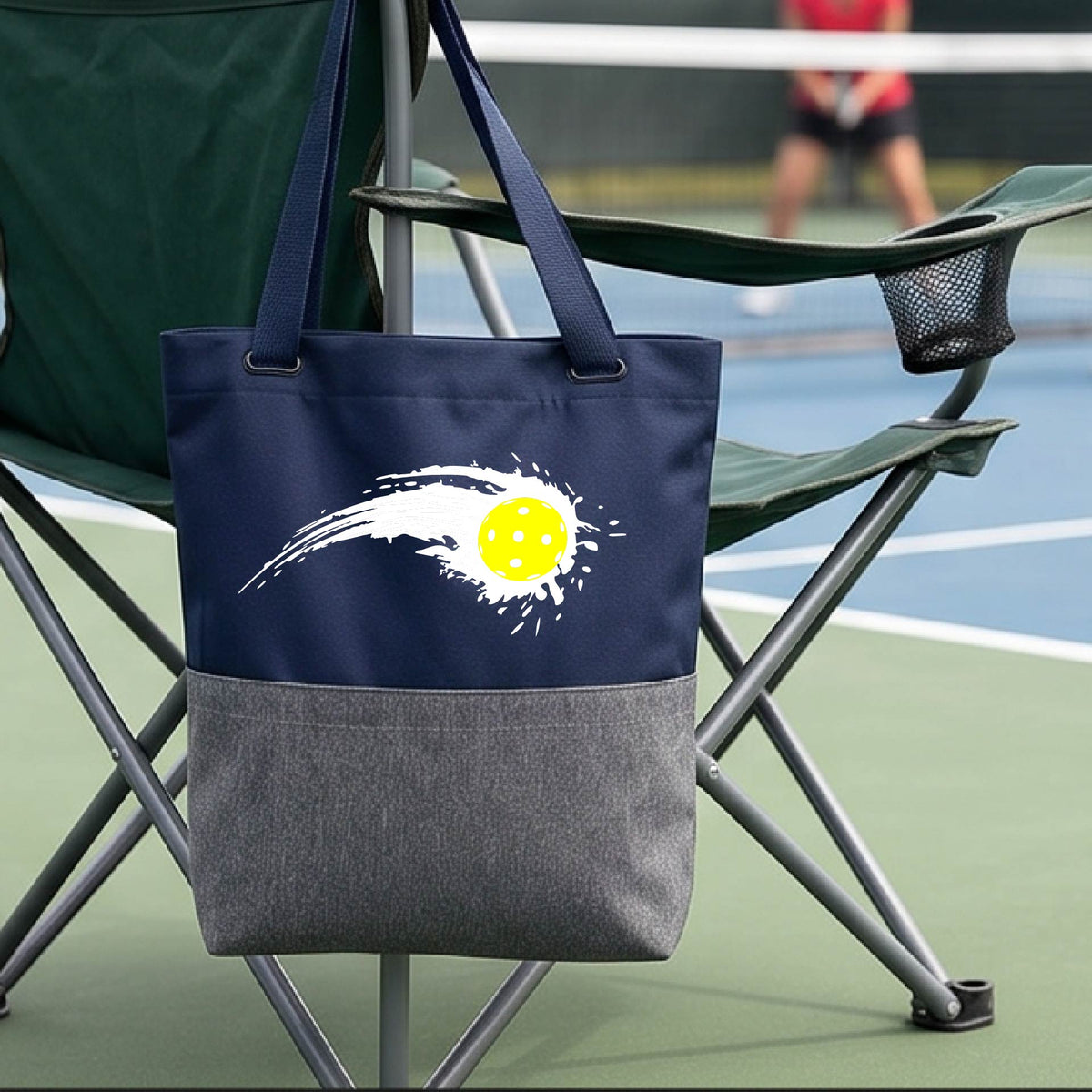A close-up of a navy blue and gray tote bag with a white pickleball graphic of a yellow pickleball with a motion trail effect, displayed in front of a pickleball court. 