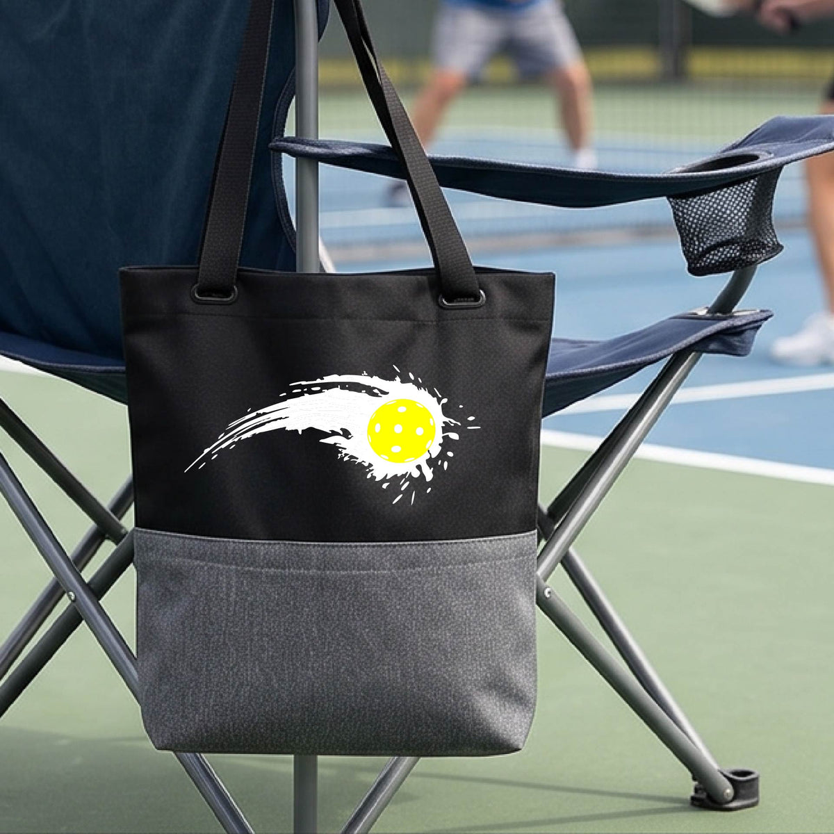 A close-up of a black and gray tote bag with a white pickleball graphic of a yellow pickleball with a motion trail effect, displayed in front of a pickleball court. 