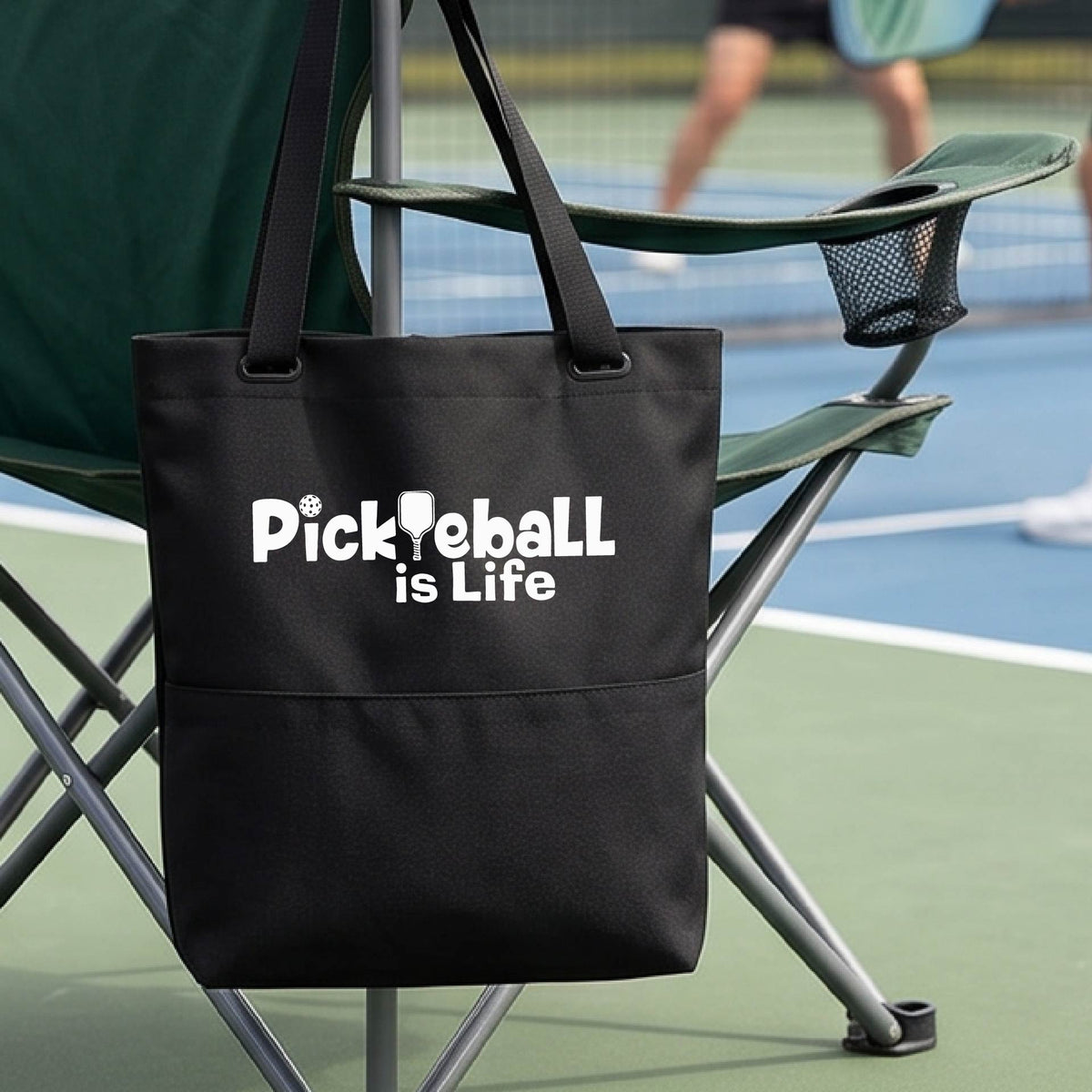 A black tote bag with the text 'Pickleball is Life' printed in white, hanging from a green folding chair on a pickleball court with a player blurred in the background.