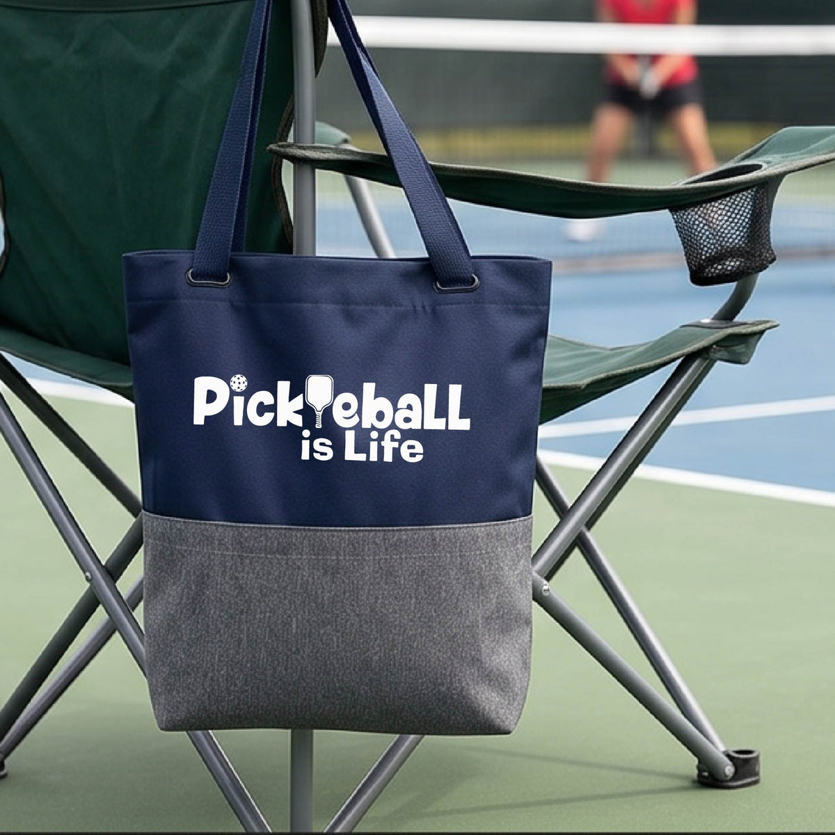 A navy blue and gray tote bag with the text 'Pickleball is Life' printed in white, hanging from a green folding chair on a pickleball court with a player blurred in the background.