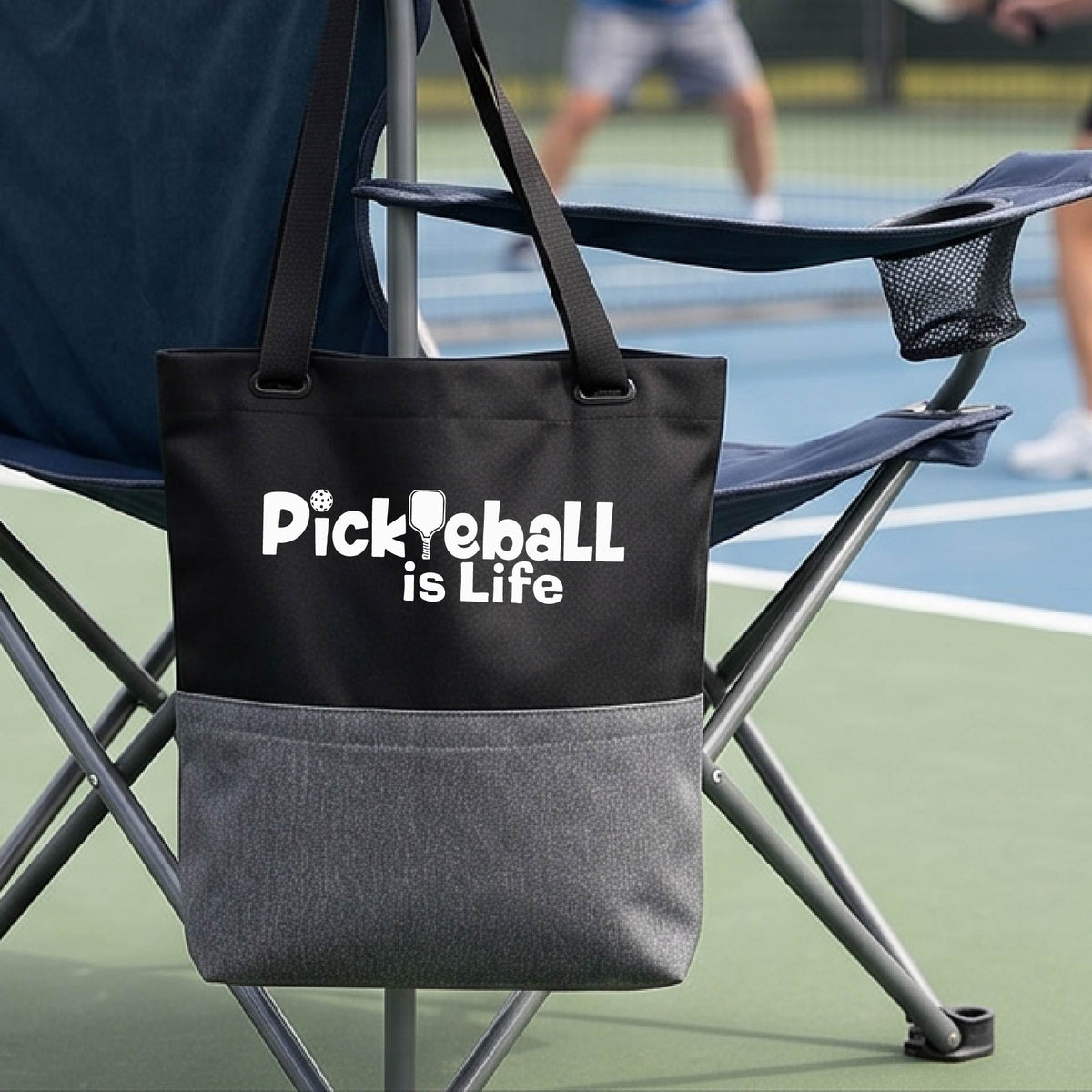 A black and gray tote bag with the text 'Pickleball is Life' printed in white, hanging from a green folding chair on a pickleball court with a player blurred in the background.