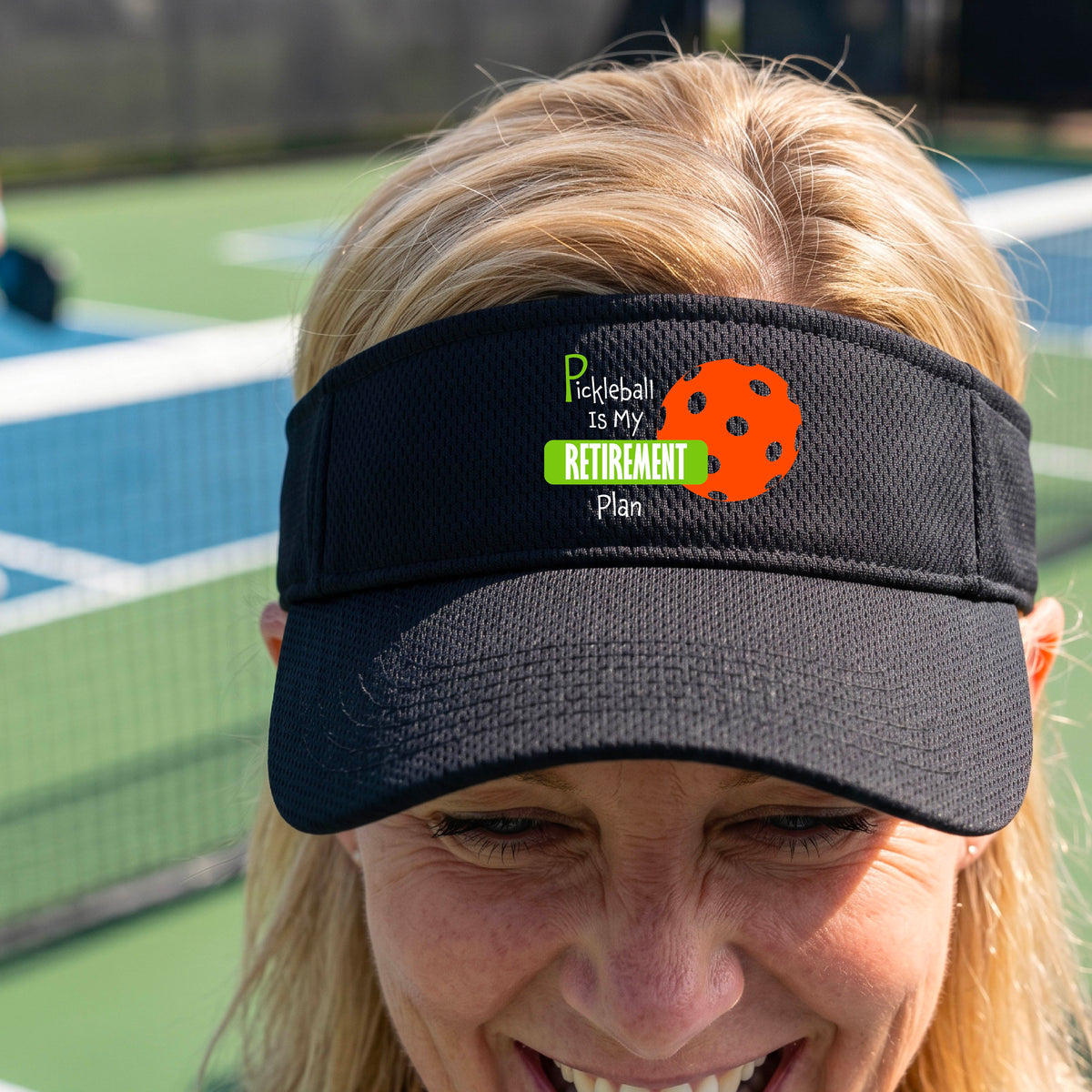 A close-up of a black visor with a custom graphic that reads 'Pickleball is my Retirement Plan' with a green and orange design, set against a blurred outdoor background.