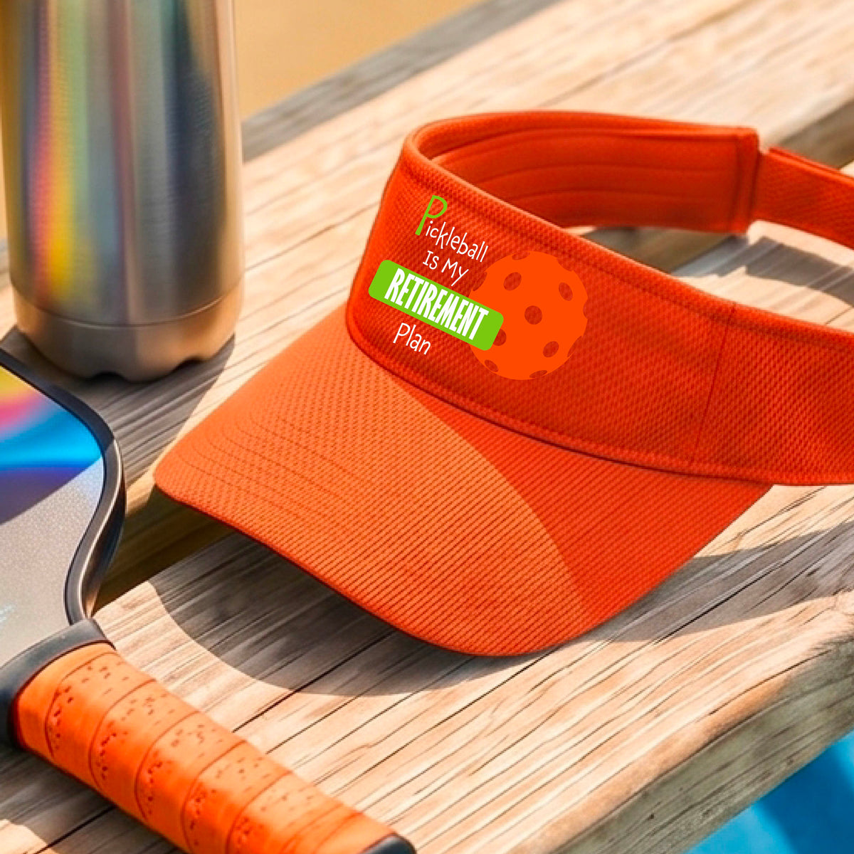 A close-up of an orange visor with a custom graphic that reads 'Pickleball is my Retirement Plan' with a green and orange design, set against a blurred outdoor background.
