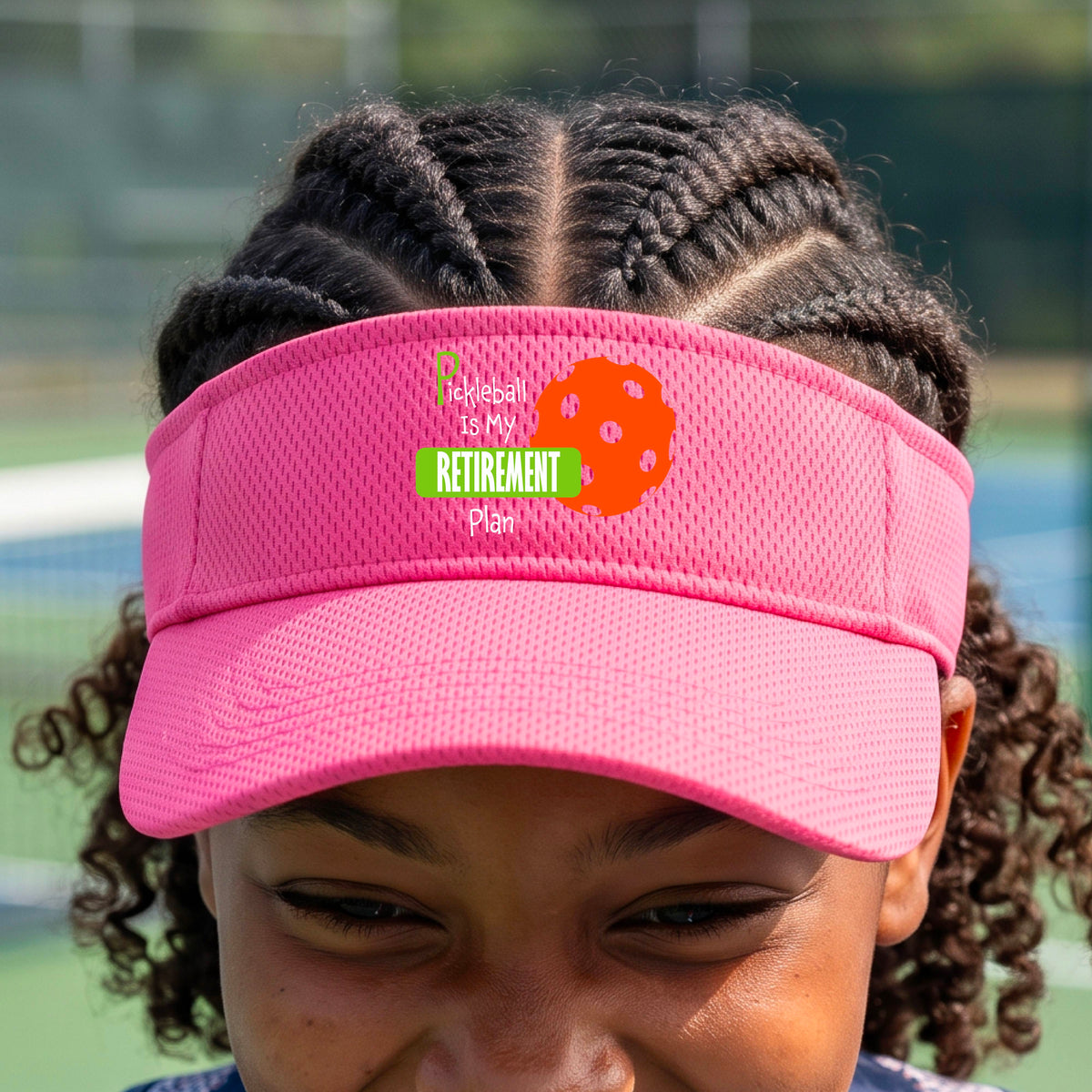 A close-up of a pink visor with a custom graphic that reads 'Pickleball is my Retirement Plan' with a green and orange design, set against a blurred outdoor background.