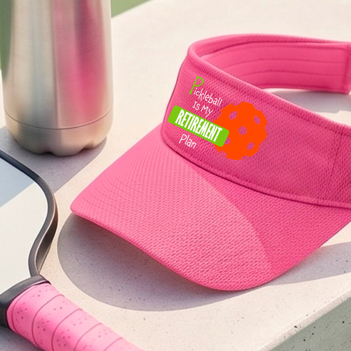 A close-up of a pink visor with a custom graphic that reads 'Pickleball is my Retirement Plan' with a green and orange design, set against a blurred outdoor background.